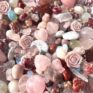 May include: A collection of various pink and clear crystals, including rose quartz hearts, carved roses, and tumbled stones. Small glass bottles filled with dried red flowers are scattered throughout the arrangement. The crystals vary in size and shape.