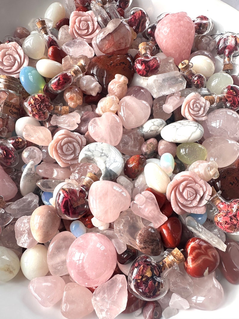 May include: A collection of various pink and clear crystals, including rose quartz hearts, carved roses, and tumbled stones. Small glass bottles filled with dried red flowers are scattered throughout the arrangement. The crystals vary in size and shape.