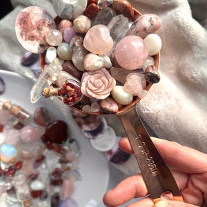 May include: A copper measuring cup filled with an assortment of polished gemstones, including rose quartz spheres, a carved rose, and a crescent moon. The cup is labeled "1 CUP 236ML". The gemstones are in shades of pink, white, and clear.
