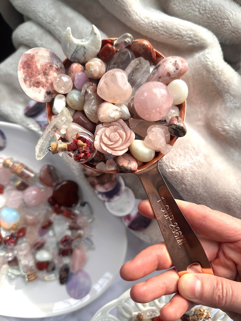 May include: A copper measuring cup filled with an assortment of polished gemstones, including rose quartz spheres, a carved rose, and a crescent moon. The cup is labeled "1 CUP 236ML". The gemstones are in shades of pink, white, and clear.
