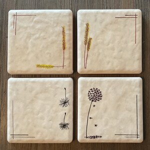 May include: Set of four square ceramic coasters with different designs. Two coasters feature a simple line drawing of a plant with brown stems and yellow leaves. The other two coasters feature a simple line drawing of a dandelion with a brown stem and black seeds.