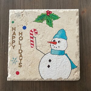 May include: Square, off-white stone coaster with a hand-drawn snowman wearing a blue hat and scarf, holding a candy cane. The words "HAPPY HOLIDAYS" are written vertically on the left side, with snowflakes and holly accents.