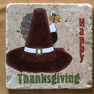 May include: A beige stone coaster with a painted image of a turkey wearing a brown pilgrim hat with a white band and a gold buckle. The words "Happy Thanksgiving" are painted on the coaster in red and green.