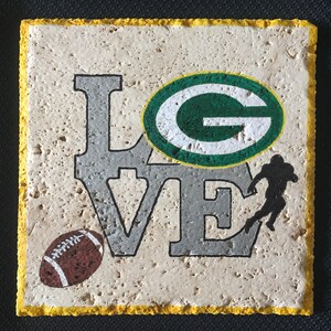 May include: A white ceramic coaster with a yellow border. The coaster features a gray "LOVE" with a green and white "G" in the center. A brown football and a black silhouette of a football player are also on the coaster.