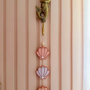 May include: A gold mermaid hook with three pink, white and peach seashells hanging from it. The seashells have a pearl-like bead in the centre.