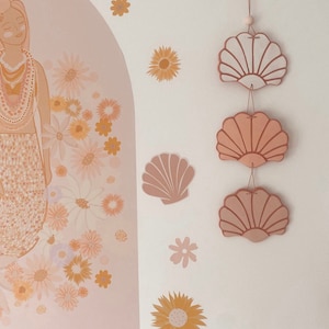 May include: A wall hanging featuring three ceramic seashells in shades of pink and white. The shells are strung together with twine and hang from a white wall.