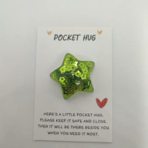 May include: A green star-shaped Pocket Hug with glitter and floral accents, presented on a white card. The card features the text "POCKET HUG" and a message about keeping it safe. A small red heart is also visible.
