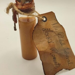 May include: A small glass bottle filled with a shimmering orange liquid. The bottle is sealed with a cork and a star-shaped stopper. A brown paper tag with handwritten text, "The Gilded Interval Locked Moments," is attached.