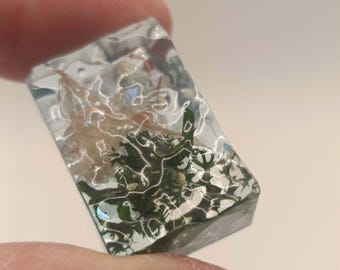 May include: A rectangular clear resin block containing embedded natural elements, including dark green moss and light-colored dried plant fibers, creating a layered, textured appearance within the transparent material.