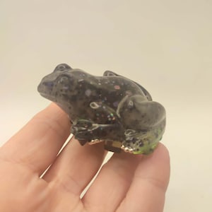 以下が含まれることがあります： イリデセントグリッターのアクセントが付いた、濃い灰色のカエルの置物。カエルは質感のある外観で、緑と銀色のディテールのある表面に置かれています。装飾品です。