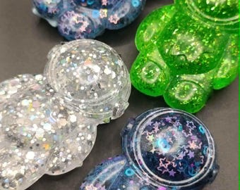 May include: Four astronaut-shaped resin figures featuring embedded glitter and star-shaped sequins. The collection includes one bright green figure, one silver glitter figure, and two dark blue figures with iridescent star accents throughout.