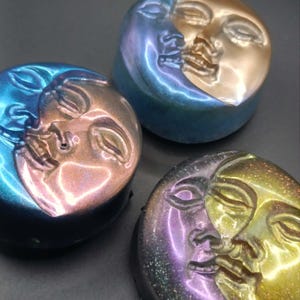 May include: Three circular objects featuring a raised relief of a face merging with a crescent moon. One object has a blue and copper finish, another is blue and gold, and the third displays a purple and yellow glittery finish with a glossy texture.