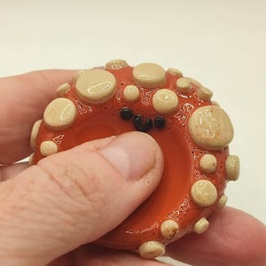 May include: A handcrafted, orange-red octopus-shaped glass bead with a smiling face and beige, rounded protrusions. The bead is held in a hand, showcasing its unique, whimsical design. This decorative item is perfect for jewelry making.