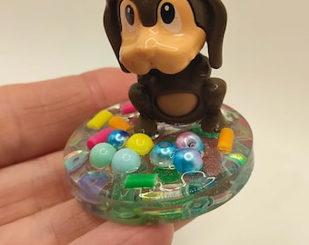 May include: A brown cartoon dog figurine with a light brown snout and ears sits atop a clear, circular base. The base is embedded with colorful beads and small, rectangular pieces. The figurine is held in a hand.
