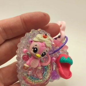 May include: A pink and white cartoon penguin charm with a cherry on its head, surrounded by a rainbow and a pink sandal. The charm is attached to a pink loop and a purple ring.
