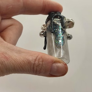 May include: A small, clear glass bottle filled with glitter and liquid. The bottle has a black top with silver chain and charm accents. The bottle is held between two fingers.
