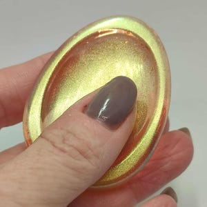 以下が含まれることがあります： 金色の内側と、キラキラと輝くマルチカラーの縁を持つ、楕円形の虹色の皿。 手で持っており、オブジェクトのサイズと色のバリエーションを示しています。