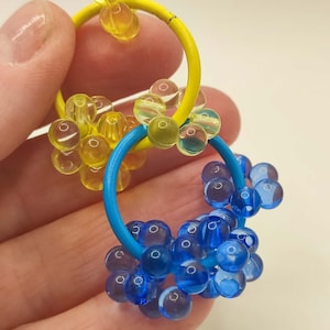 May include: A pair of handmade earrings featuring a yellow and blue hoop design. Each hoop is adorned with clusters of transparent, spherical beads in shades of yellow and blue. The earrings are held against a light background.