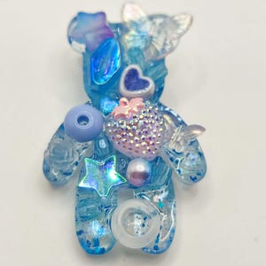 Könnte beinhalten: Blauer Bären-förmiger Broschenanhänger aus Harz mit Glitzer und verschiedenen Verzierungen. Er hat ein Herz, einen Stern, einen Schmetterling und andere dekorative Elemente in Blau-, Rosa- und transparenten Farbtönen. Ein einzigartiges, handgefertigtes Accessoire.
