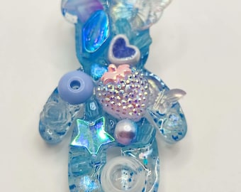 May include: A blue resin bear-shaped brooch with glitter and various embellishments. It features a heart, star, butterfly, and other decorative elements in shades of blue, pink, and clear. The brooch is a unique, handcrafted accessory.