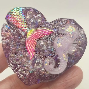 May include: A heart-shaped resin piece featuring a mermaid tail and a seahorse. The mermaid tail has iridescent scales and a pink and yellow fin. The seahorse is light purple. The resin is purple with clear bubbles.