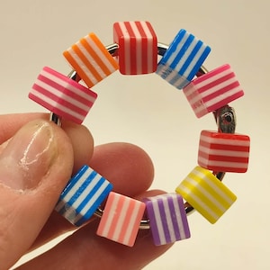 以下が含まれることがあります： オレンジ、赤、青、ピンク、黄色、紫など、さまざまな色のストライプ模様の小さな立方体ビーズのカラフルなリング。 ビーズは銀色の金属リングに取り付けられており、遊び心のある鮮やかなアクセサリーになっています。