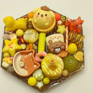 May include: A hexagonal resin piece with miniature food-themed charms. It features a yellow dog character, a fox, stars, donuts, and sweets in yellow, orange, and brown. The base is a translucent brown. The charms are arranged in a decorative pattern.