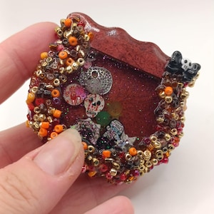 Könnte beinhalten: Ein Resin-Shaker-Anhänger mit einem tiefroten, glitzernden Zentrum, das mit Totenkopf-, Handabdruck- und kreisförmigen Charms gefüllt ist. Der Rand ist mit einer dichten Ansammlung von orangefarbenen, braunen und goldenen Perlen sowie einer kleinen schwarzen Schleife verziert.