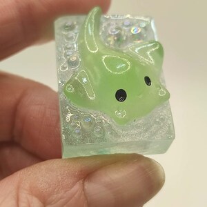 May include: A small, rectangular resin charm featuring a green manta ray with black eyes. The charm is set in a clear, glittery base with bubble-like details, creating a whimsical, underwater effect. The manta ray has a cute, cartoonish design.