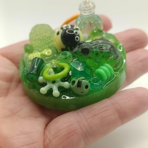 May include: A circular green resin charm featuring an embedded collection of miniature objects including a dark green frog, a small black and white creature, a clear glass-like bottle, a white star-shaped piece, and various green beads.