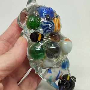Könnte beinhalten: Eine klare Glasskulptur, gefüllt mit bunten Murmeln verschiedener Größen und Muster. Die Murmeln umfassen Farbtöne von Blau, Grün, Orange und Weiß, eingeschlossen in einer klaren, amorphen Form. Die Skulptur wird in einer Hand gehalten.