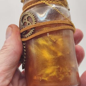 May include: A clear glass bottle filled with a shimmering gold liquid. The bottle is adorned with brown leather straps, a gold chain, and a bronze gear. The top of the bottle has a metallic gold wax seal. The overall aesthetic is steampunk.