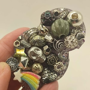 May include: A handmade resin brooch with various embellishments. The brooch includes a skull, stars, beads, and a rainbow. The colour palette includes silver, gold, green, and purple. The brooch is held in a hand.