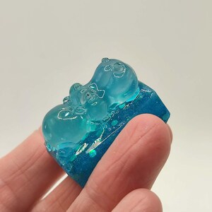 May include: A clear, blue resin sculpture featuring two hippo figures. The hippos are a lighter shade of blue and appear to be embracing. The base is a darker blue with glitter accents. The sculpture is held in a hand.