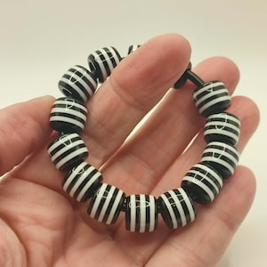 May include: A black and white striped bead bracelet. The bracelet features cylindrical beads with alternating black and white stripes. The beads are strung together, forming a circular shape. The bracelet is held in a hand against a white background.