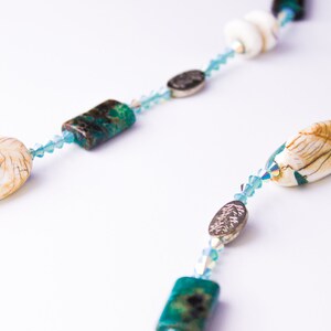 May include: A necklace featuring a strand of light blue crystal beads, interspersed with white and tan shell-shaped beads, dark teal rectangular beads, and silver-toned oval beads. The necklace is displayed against a white background.