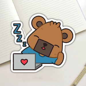May include: A sticker featuring a cartoon bear wearing a blue shirt, sleeping in front of a laptop with a heart on the screen. The sticker has a white border and the word "Z" repeated three times above the bear's head.
