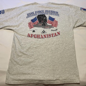 May include: A light gray t-shirt featuring a graphic of a black panther, American flags, and the text "TASK FORCE PANTHER OPERATION ENDURING FREEDOM AFGHANISTAN." The shirt also has patches on the sleeves.