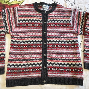 May include: A patterned cardigan with a button-down front, featuring a design in red, black, grey, and cream. The sweater has a round neck and long sleeves. The label reads "14001:001:01".