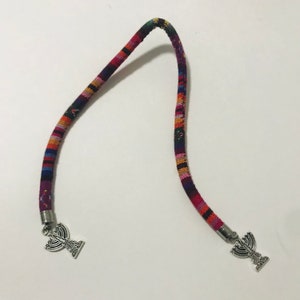 May include: A colorful, woven bookmark with silver menorah charms at each end. The bookmark is made of a thick, braided cord.