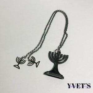 Puede incluir: Un collar de metal negro con un colgante de menorá. El colgante tiene la palabra "Jerusalén" inscrita en la base. El collar también tiene dos pequeños encantos de menorá unidos a la cadena.