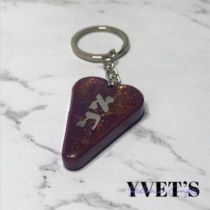 Pode incluir: Um chaveiro de resina em forma de coração roxo com acabamento de glitter prateado e uma inscrição de metal prateado da palavra hebraica "Chai".