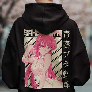 Puede incluir: Sudadera con capucha negra con un personaje de anime rosa, orejas de conejo y pelo rosa. El diseño incluye texto japonés y la palabra "SAK". El fondo es una escena borrosa al aire libre.
