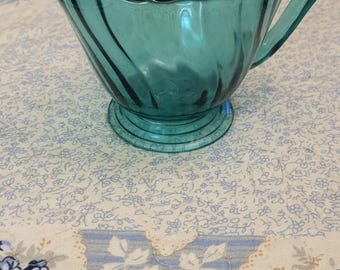 May include: A teal blue glass creamer with a fluted design and a handle. The creamer is sitting on a floral patterned tablecloth.