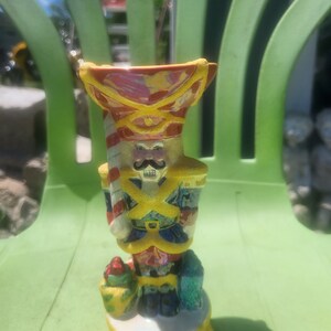 May include: A ceramic nutcracker figurine with a red and yellow hat, a white and red striped candy cane, and a yellow and blue uniform. The figurine is holding a bag of gifts and is standing on a yellow base with a star.