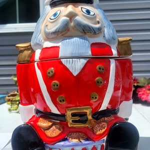 Puede incluir: Un tarro de galletas de cerámica con forma de soldado Cascanueces. El tarro es predominantemente rojo con detalles blancos, botones dorados y una hebilla dorada. El Cascanueces tiene un sombrero rojo, ojos azules y un bigote gris. La base es un tambor.