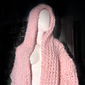 May include: A light pink, chunky knit hooded cardigan. The open-front sweater is displayed on a mannequin. The thick knit has a textured appearance. The hood is large and drapes over the shoulders.