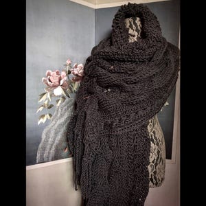 Puede incluir: Chal de punto gris oscuro con un diseño texturizado, drapeado sobre un maniquí. El chal presenta un cuello alto y bordes con flecos. Se ven pequeños alfileres decorativos. El fondo incluye una pintura floral.