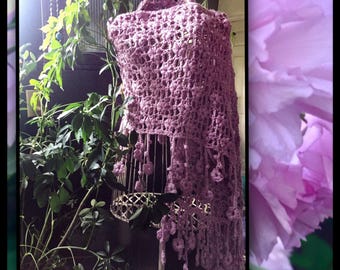 May include: A mauve crochet shawl on a wire dress form. The open weave design has a fringe with small, flower-like accents. Green plants and a large, blurred pink flower surround the shawl. The shawl is approximately 150cm long.