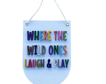 May include: A light blue wooden sign with the words "WHERE THE WILD ONES LAUGH & PLAY" in colorful, raised letters. The sign is suspended by a white string and a wooden ring.